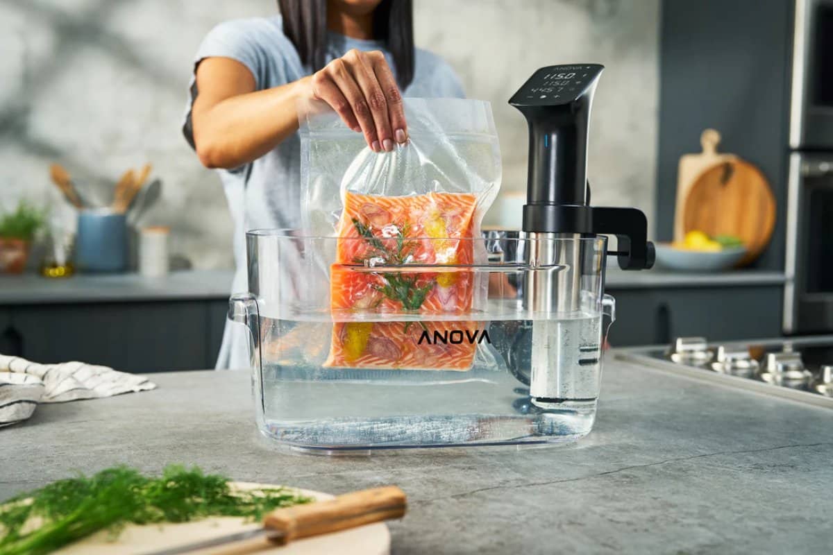 A person adds a vacuum-sealed bag of salmon to a water bath with an Anova sous vide precision cooker on a kitchen counter.