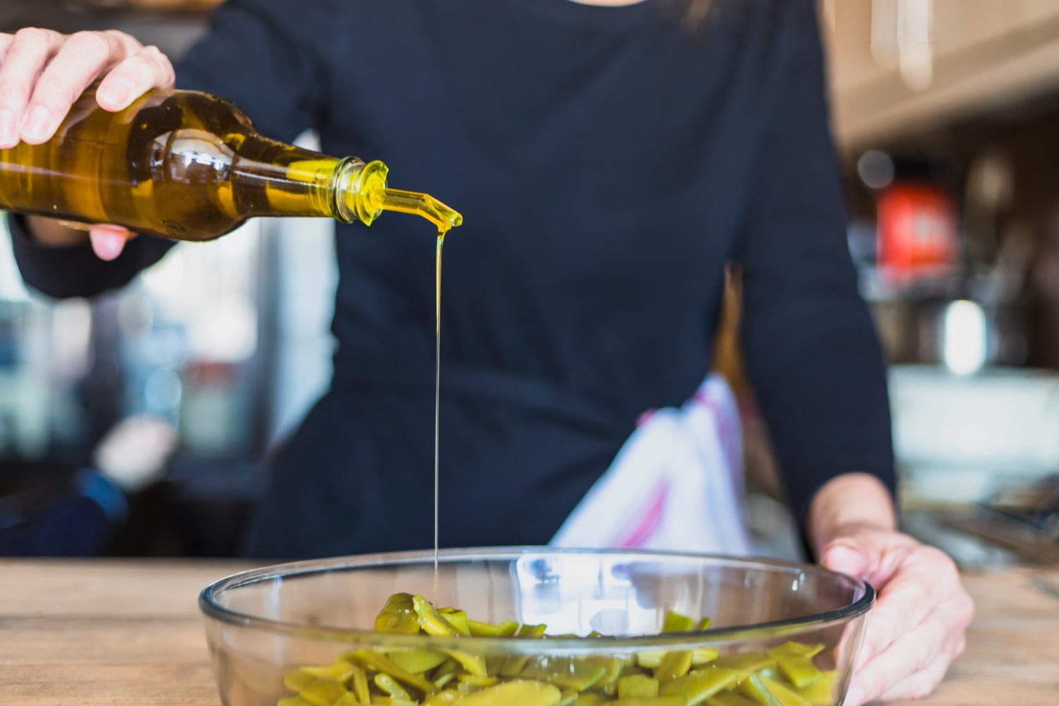 A person in a black shirt pours olive oil from a bottle into a bowl of green vegetables, preparing a fresh meal.
