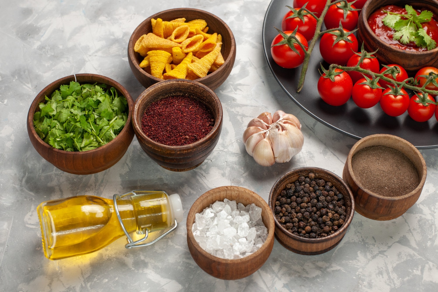 An array of fresh ingredients: vibrant cherry tomatoes, bowls of herbs and spices, a bottle of oil, and crunchy snacks on a marble surface.