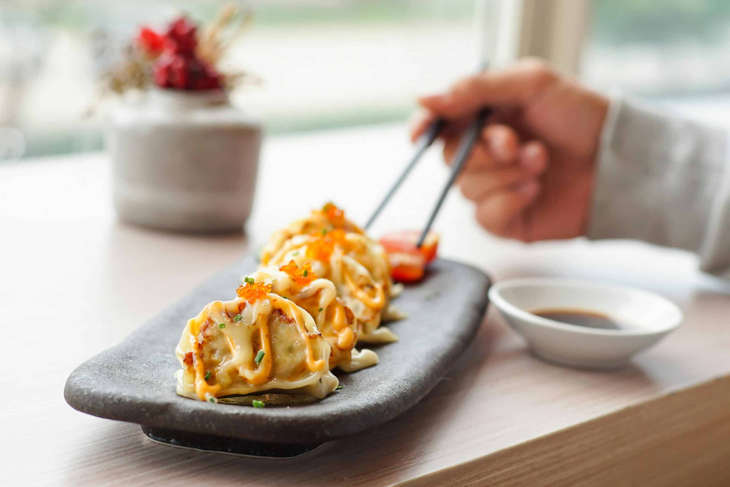 A hand using chopsticks to pick up doused dumplings, garnished with orange roe, on a stone plate, with soy sauce on the side.