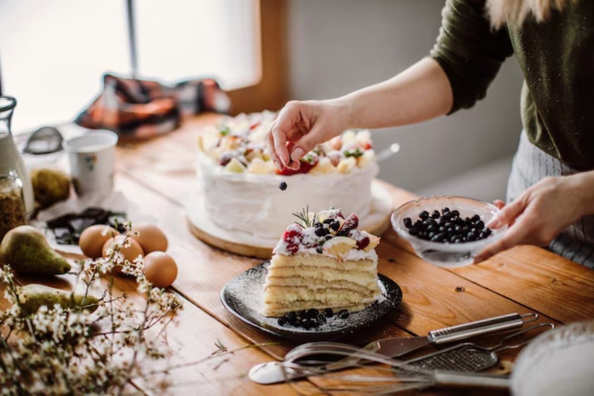 A person decorates a layered fruit cake with fresh berries, surrounded by ingredients like eggs, pears, and baking tools on a wooden table.