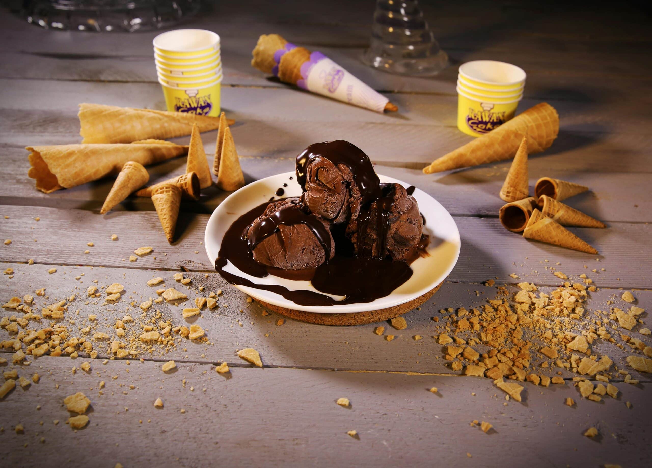 A plate of chocolate ice cream drizzled with chocolate sauce, surrounded by various ice cream cones and scattered crumbs on a wooden surface.