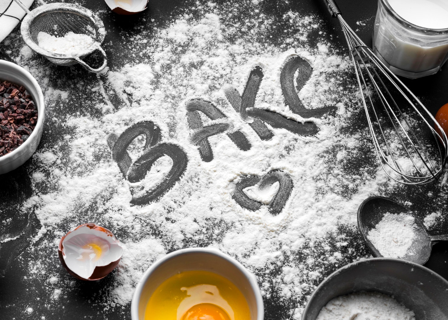 A baking surface scattered with flour, featuring the word BAKE and a heart, surrounded by eggs, a whisk, and cocoa nibs.