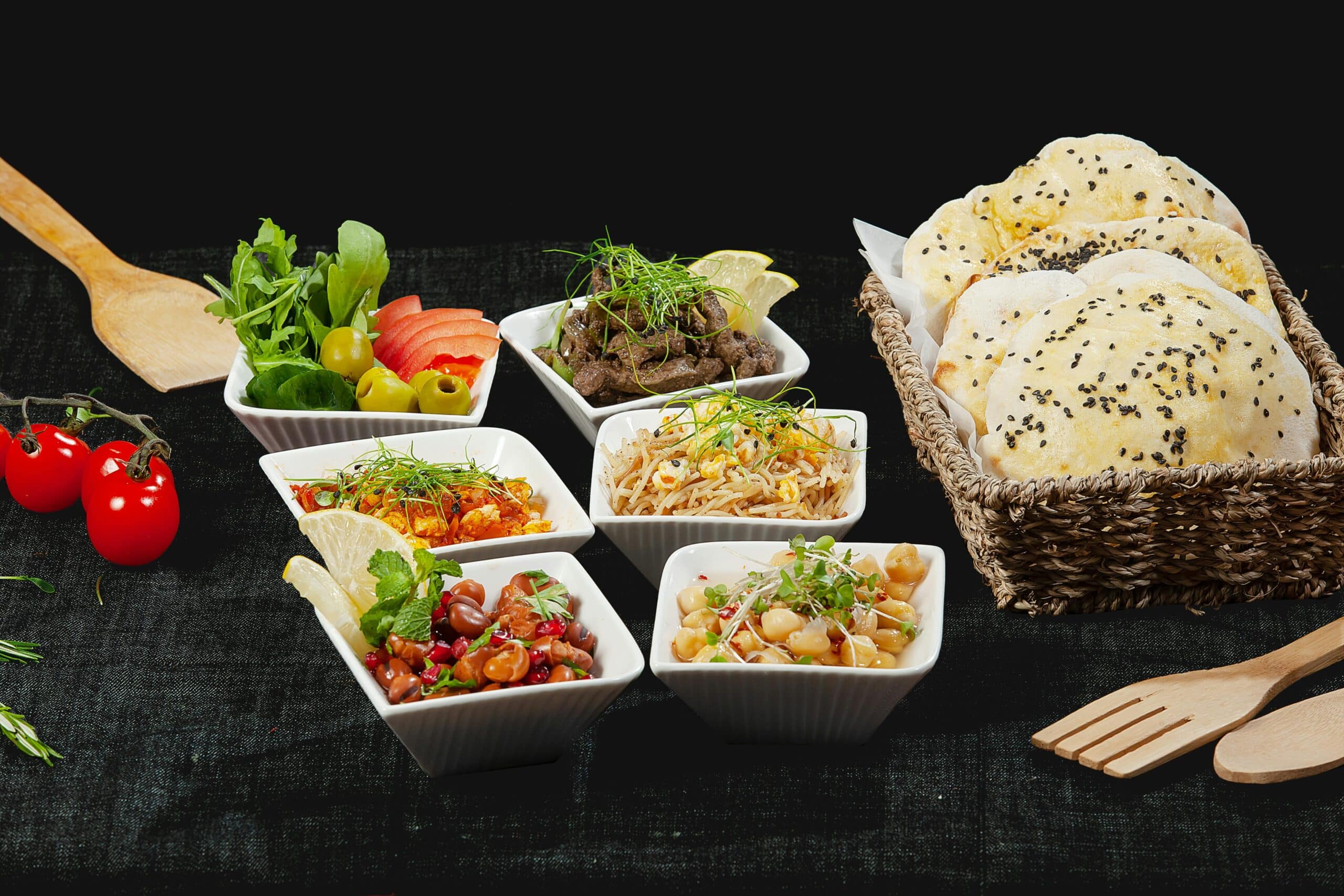 A spread of vibrant dishes, including salads, meats, and grains, served in white bowls with fresh bread in a basket alongside.