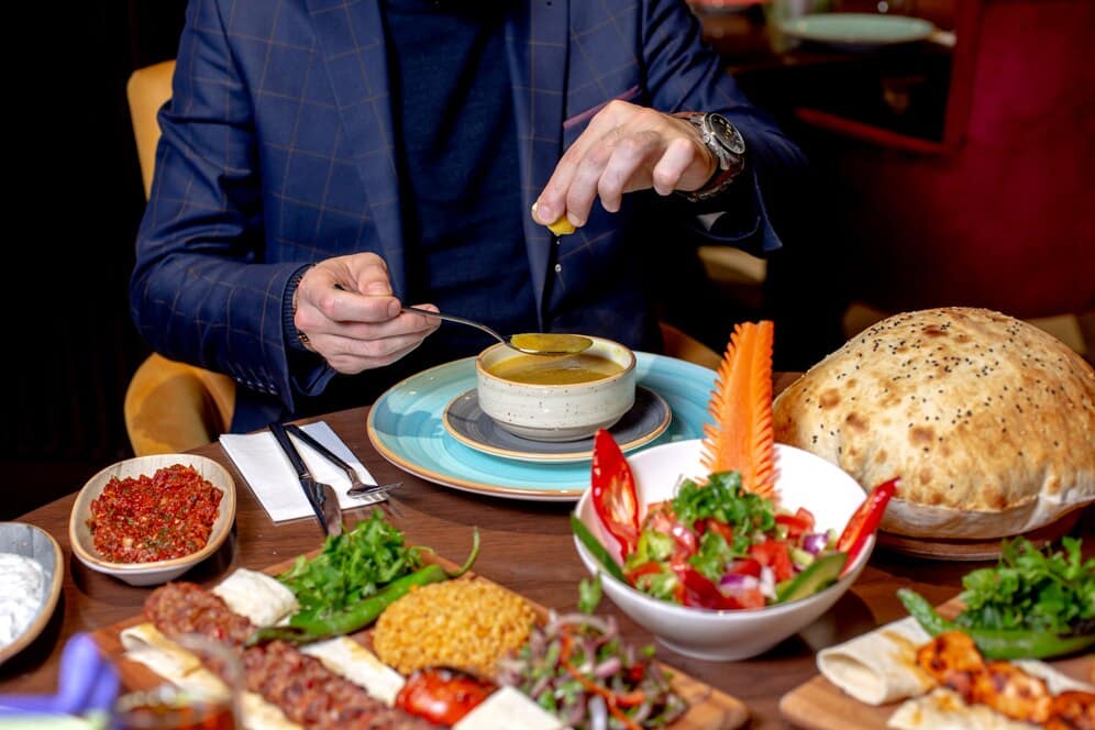 A man in a suit delicately adds lemon to a bowl of soup, surrounded by a colorful spread of salads, bread, and condiments on a dining table.