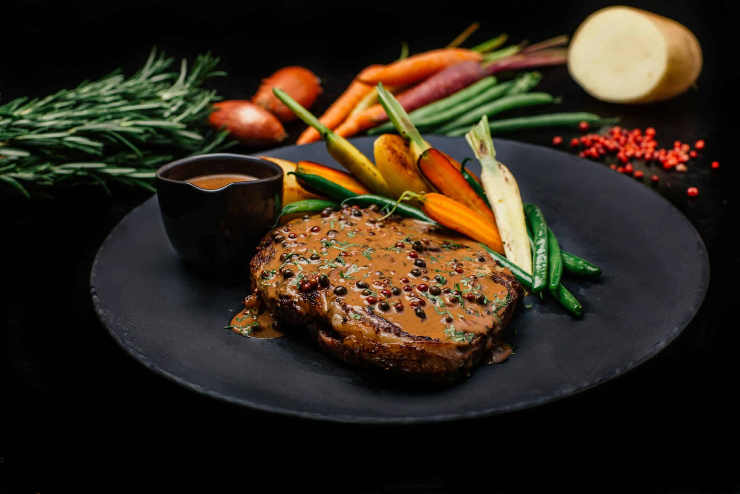 A gourmet steak covered in pepper sauce, garnished with herbs, alongside colorful roasted vegetables on a black plate.