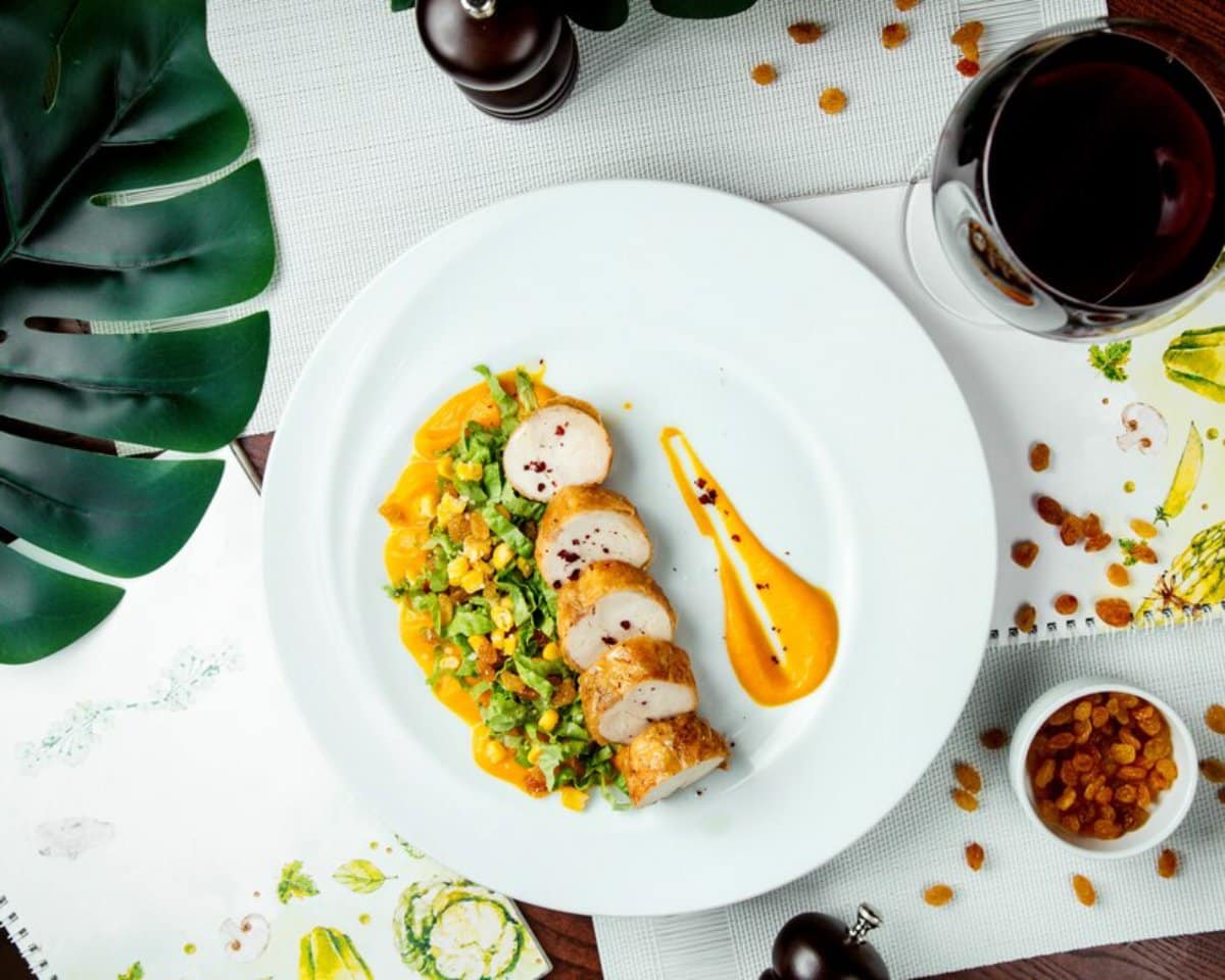 A beautifully plated dish of sliced chicken atop a salad with yellow sauce, accompanied by red wine and decorative greens.