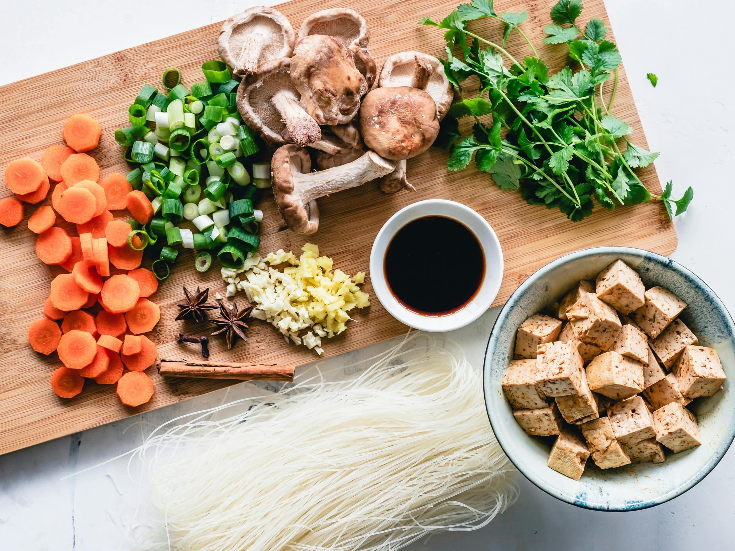 A cutting board displays vibrant sliced carrots, green onions, shiitake mushrooms, ginger, spices, tofu cubes, vermicelli noodles, and soy sauce.