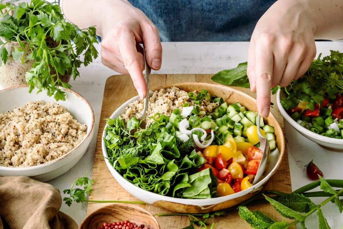A person mixes a vibrant salad with fresh greens, quinoa, tomatoes, and herbs, preparing a healthy meal in a rustic kitchen.