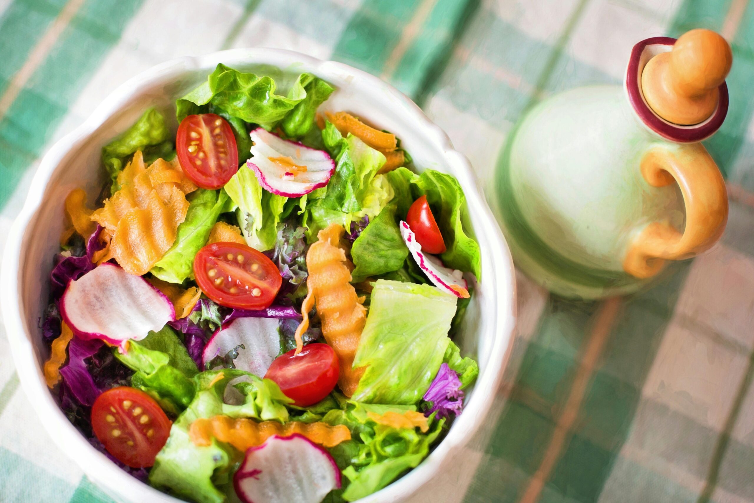A colorful salad bowl filled with greens, cherry tomatoes, radishes, and orange carrots, beside a decorative oil dispenser.