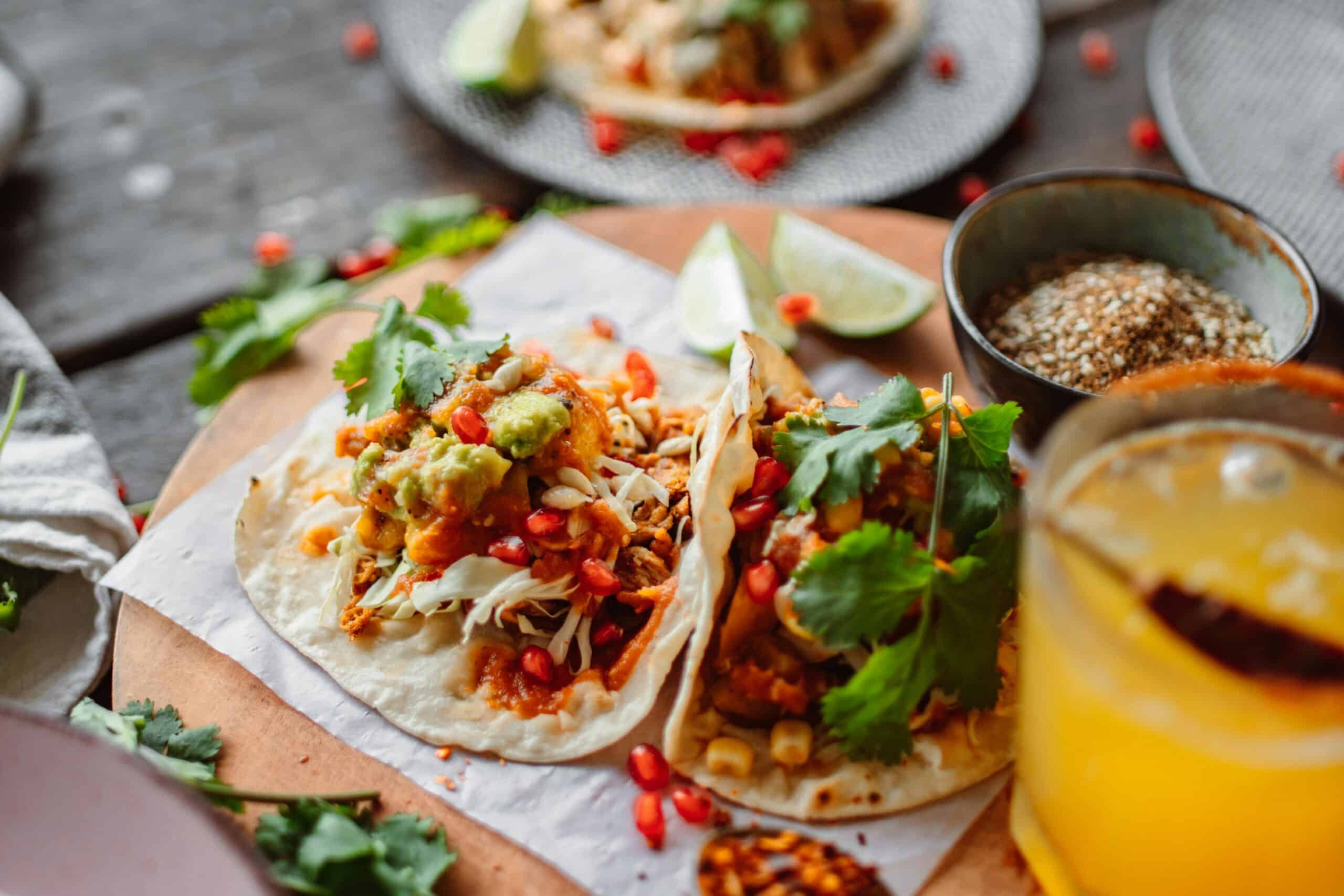 Two vibrant tacos filled with toppings like guacamole, cabbage, and pomegranate seeds, garnished with cilantro and accompanied by lime wedges.