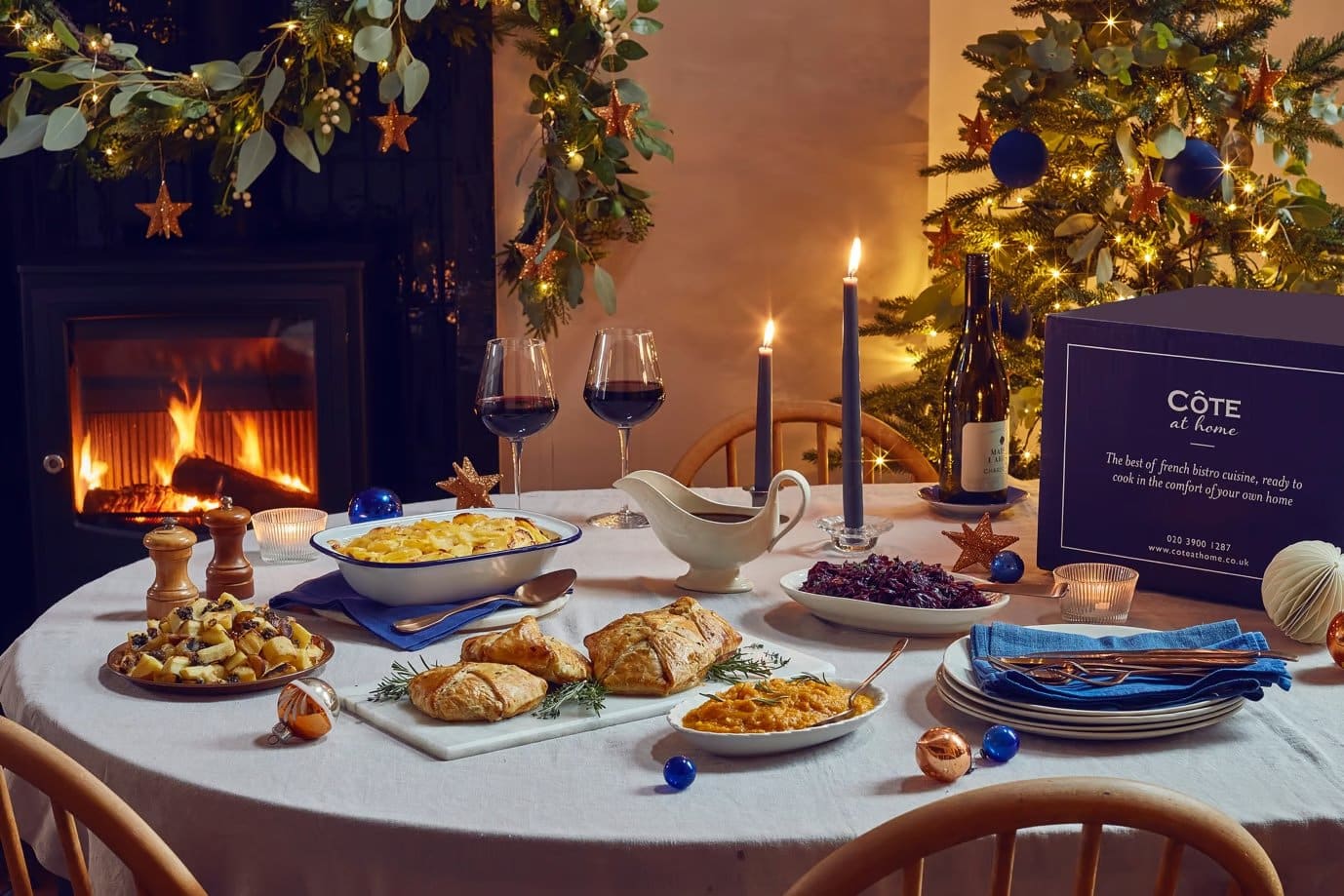 A festive dining table set with gourmet dishes, wine glasses, and candles, surrounded by holiday decorations and a glowing fireplace.