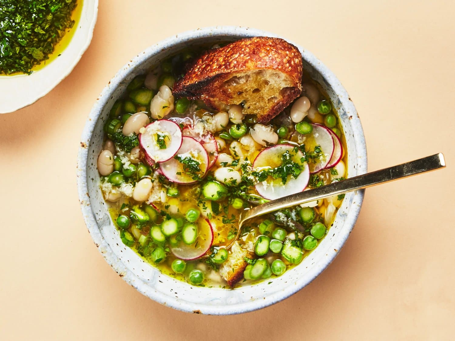 A bowl of vibrant soup with white beans, green peas, radishes, and herbs, garnished with crusty bread, set against a soft peach backdrop.