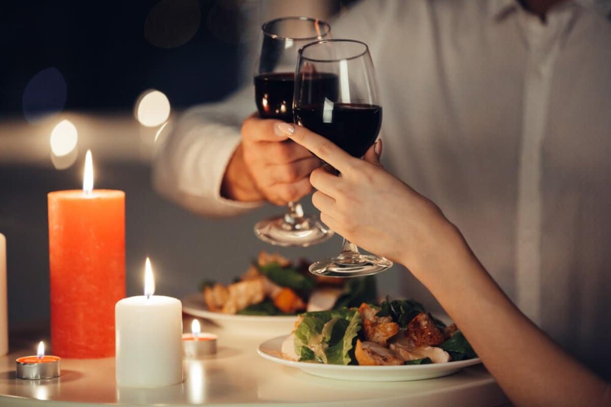 A romantic dinner scene with two hands clinking wine glasses, surrounded by candlelight, plates of food, and a cozy atmosphere.
