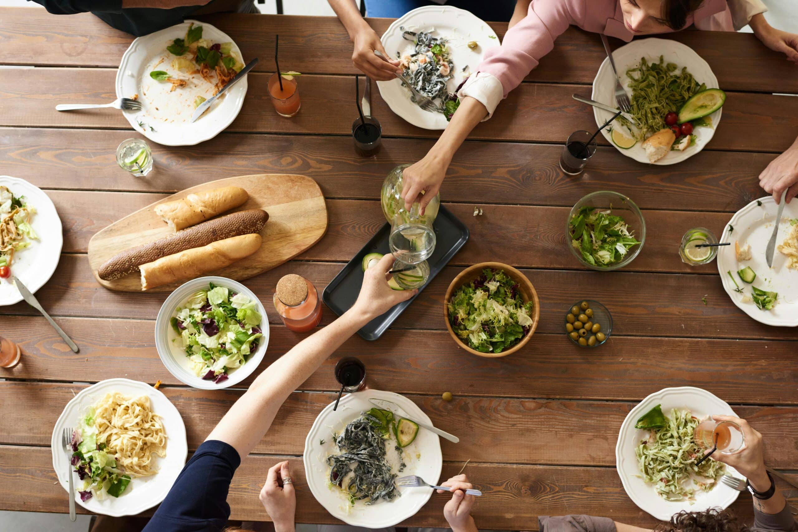 Aerial view of a wooden dining table with people enjoying various pasta dishes, salads, and drinks, sharing a lively meal.