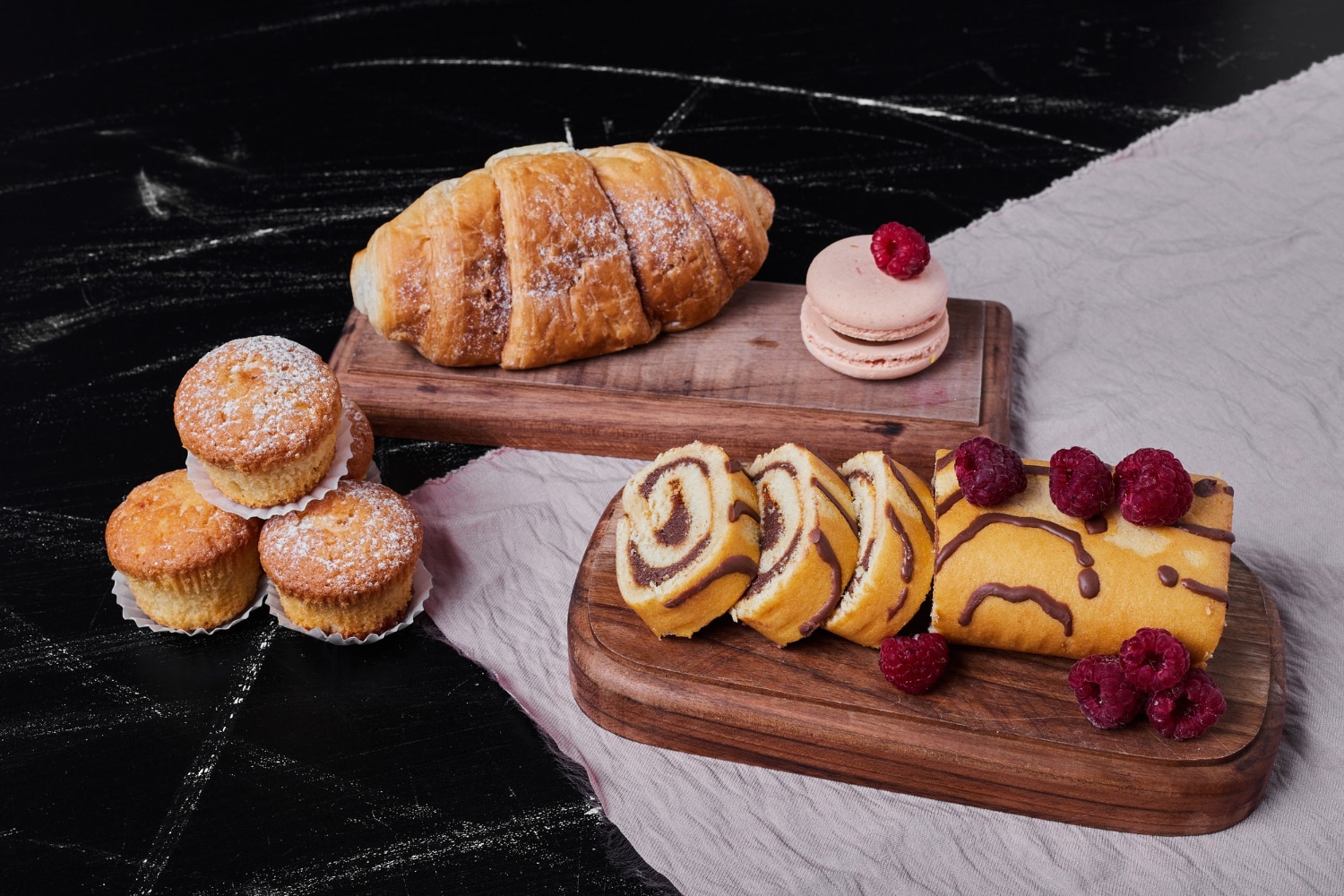 An assortment of baked goods: a croissant, macarons, cupcakes, and a chocolate swirl roll, garnished with fresh raspberries.
