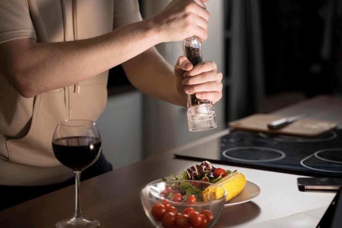 A person in a beige hoodie grinds pepper over a fresh salad with a glass of red wine nearby, all set on a kitchen table.