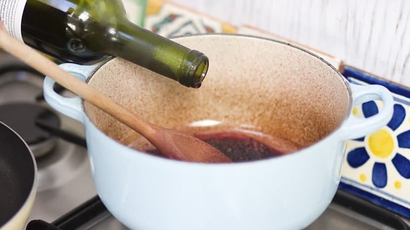 A green wine bottle being poured into a brown ceramic pot, with a wooden spoon inside, on a stove top.