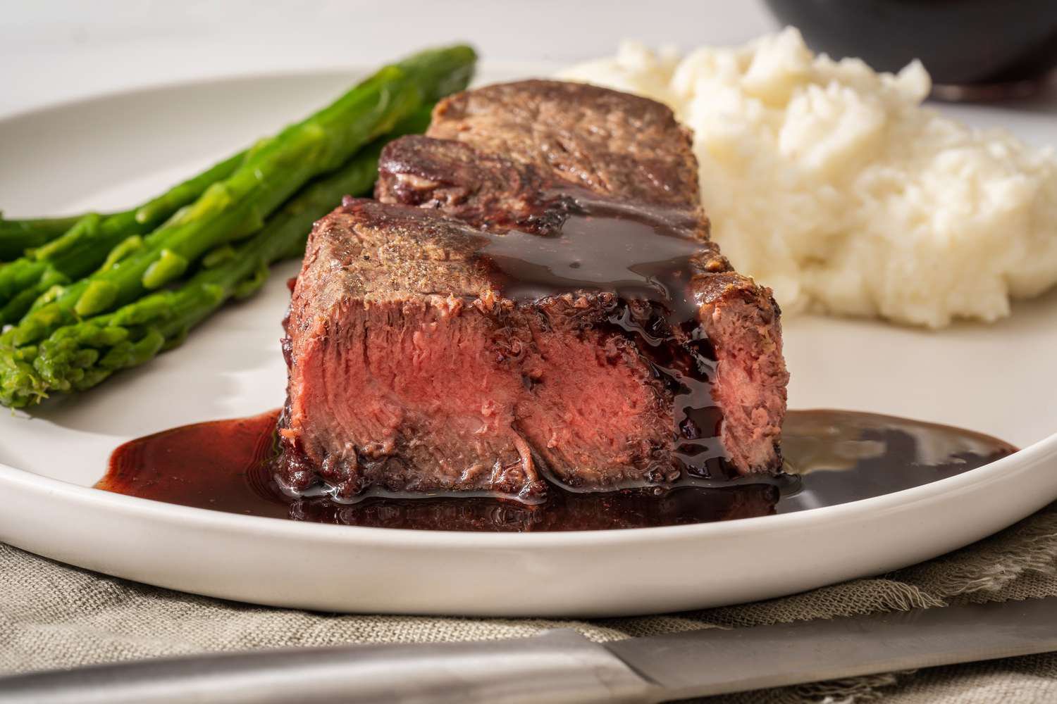 A perfectly cooked beef steak drizzled with sauce, accompanied by asparagus and creamy mashed potatoes on a white plate.