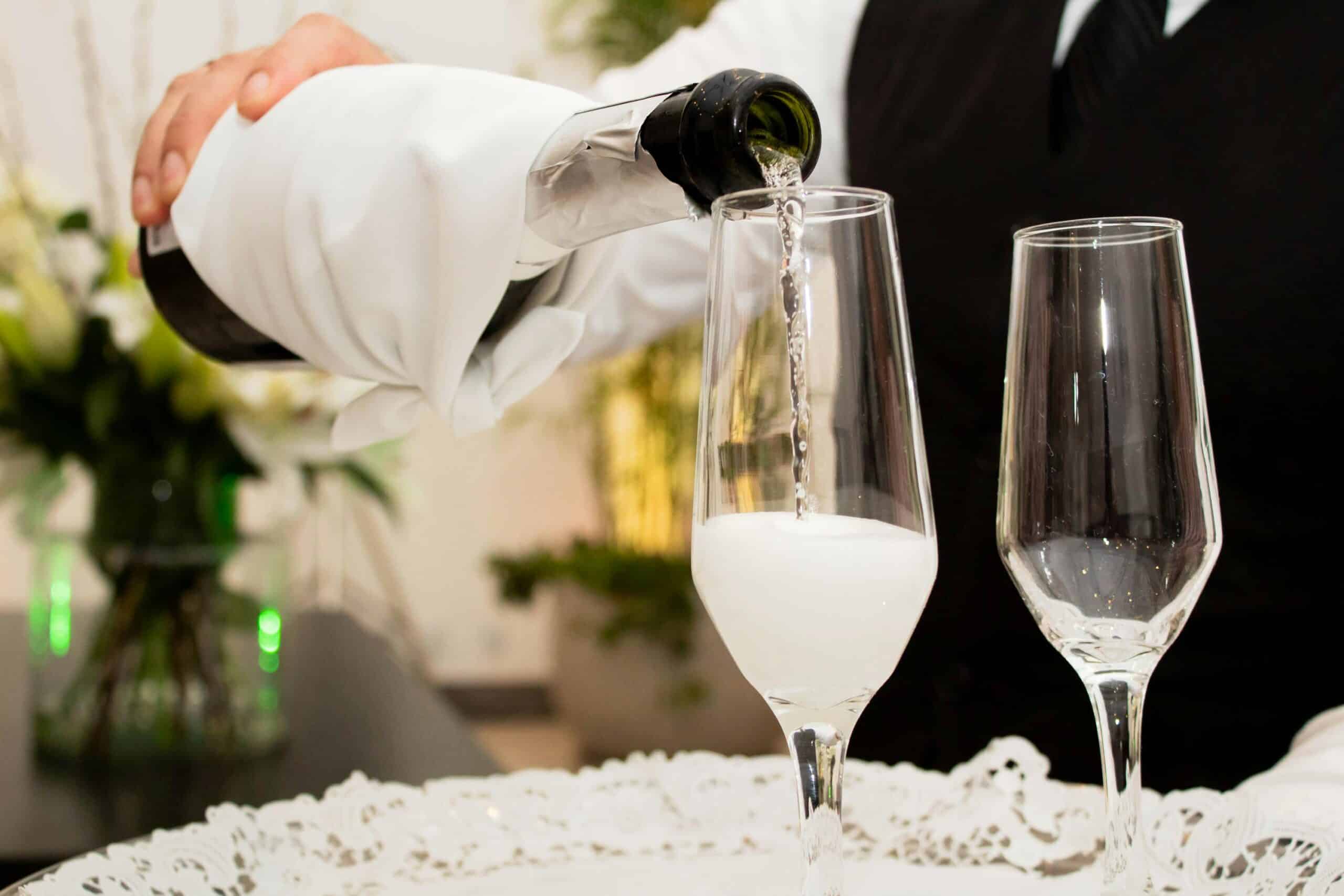 A waiter pours sparkling wine from a bottle into a glass, set on an elegant lace-trimmed tray amidst a floral background.
