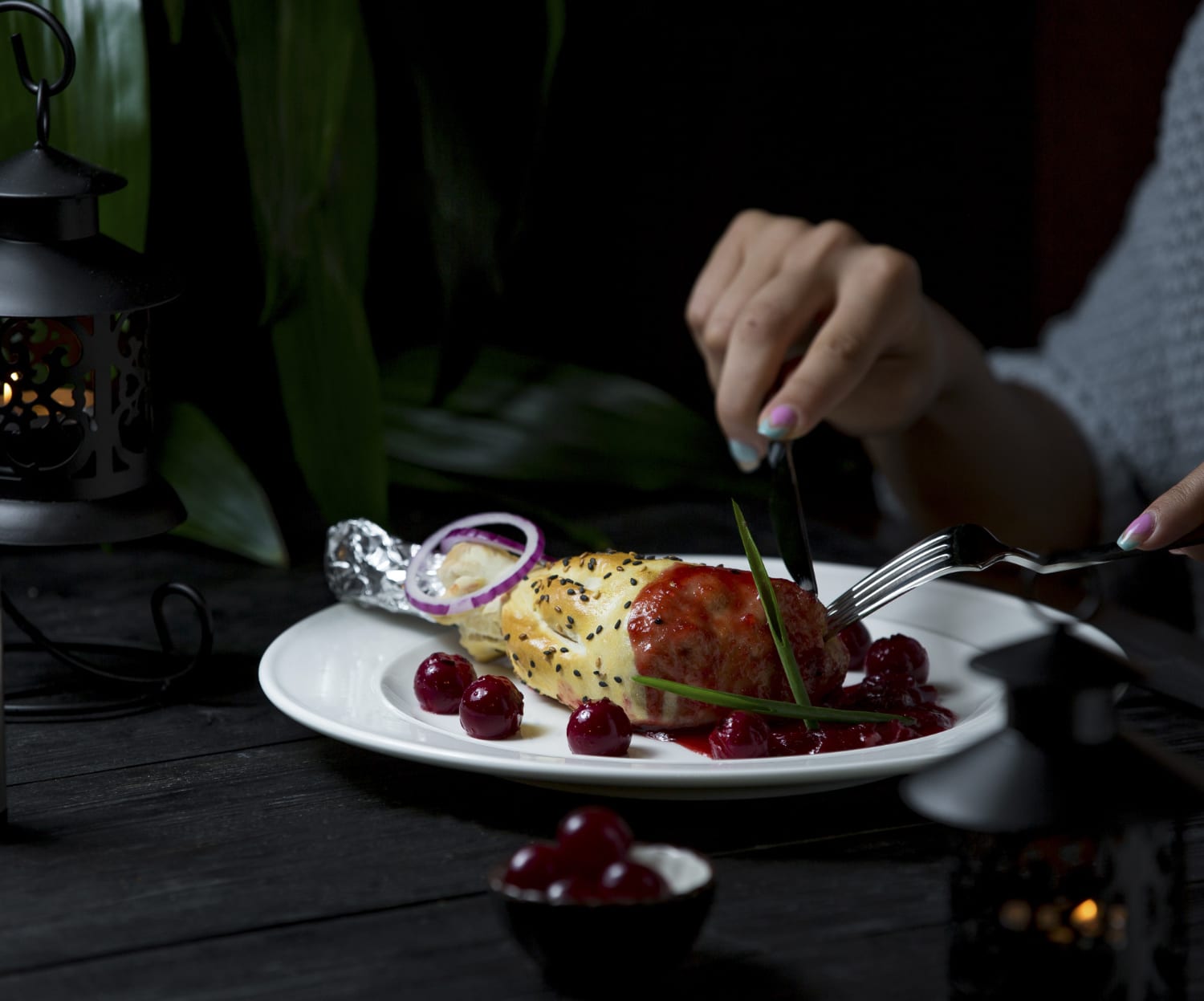 A close-up of a hand using a fork and knife on a gourmet dish with sauce, garnished herbs, and cherries, with lanterns in the background.
