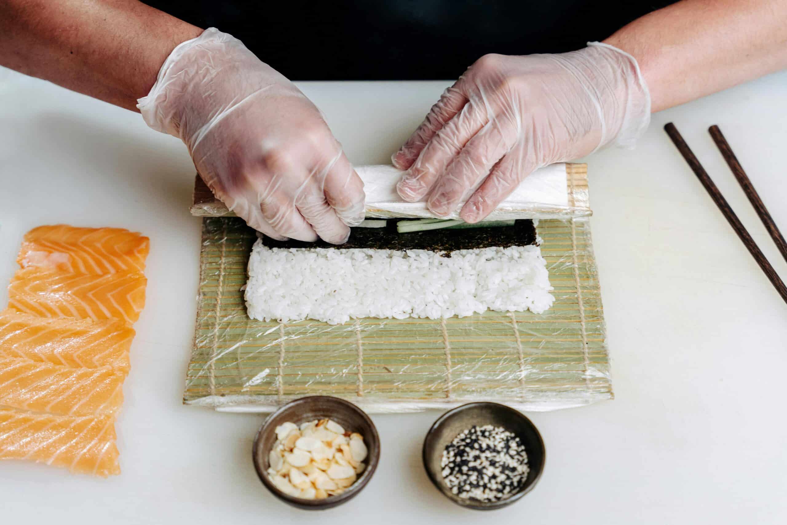 A chef in gloves rolls sushi with rice, seaweed, and cucumber, with salmon slices and bowls of toppings in the background.