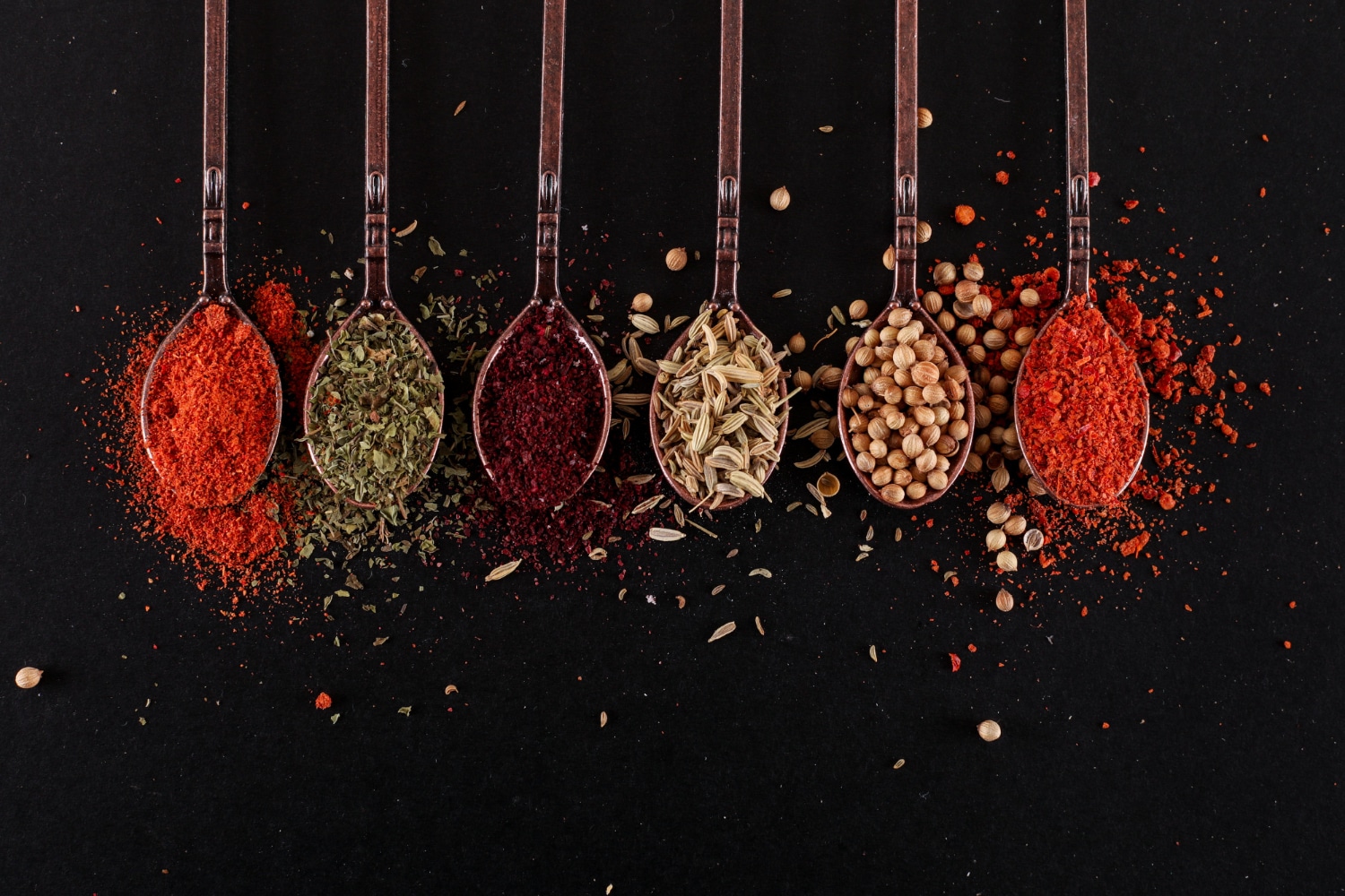 A selection of colorful spices and herbs poured into vintage spoons, arranged against a dark background.