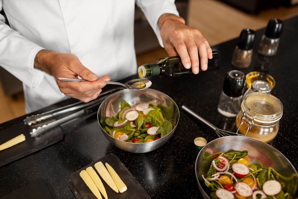 A chef drizzles olive oil onto a fresh salad with spinach, radishes, and cherry tomatoes, surrounded by seasonings on a kitchen counter.