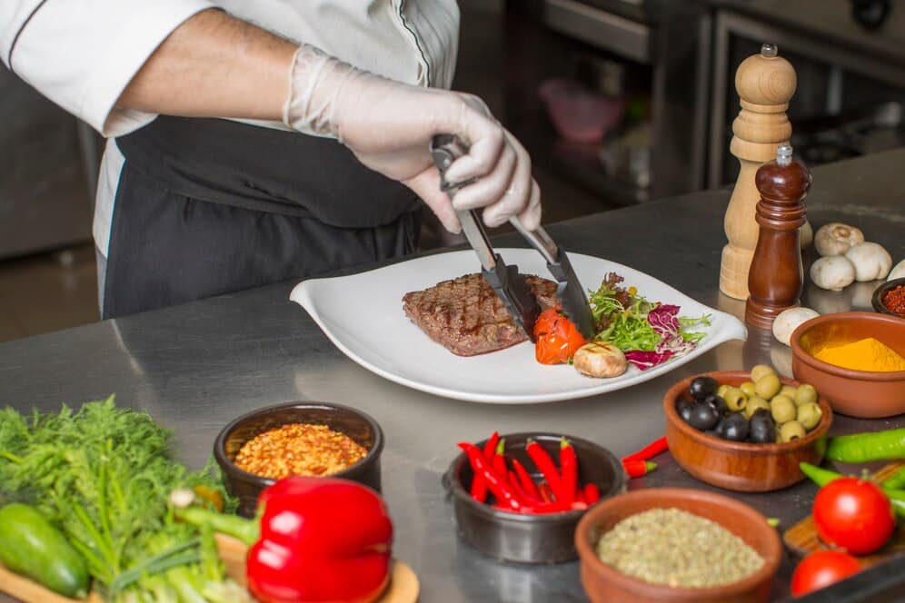 A chef in a white coat uses tongs to place grilled meat and vegetables on a white plate, surrounded by colorful fresh ingredients.