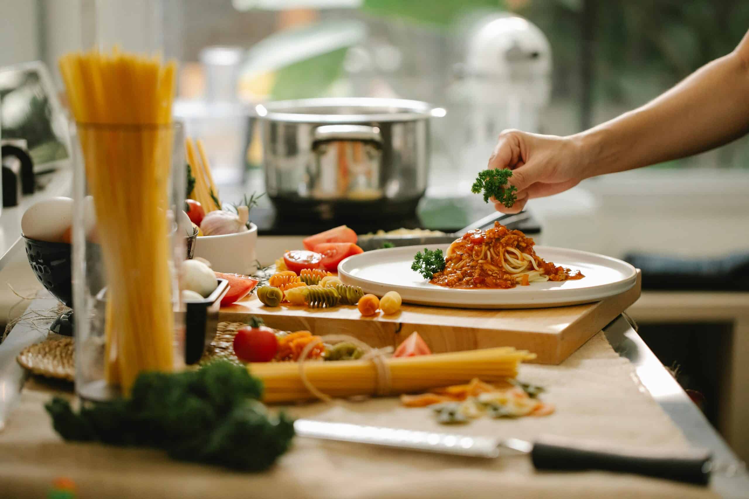 A hand garnishes a plate of spaghetti with marinara sauce and parsley, surrounded by fresh ingredients and pasta on a kitchen counter.