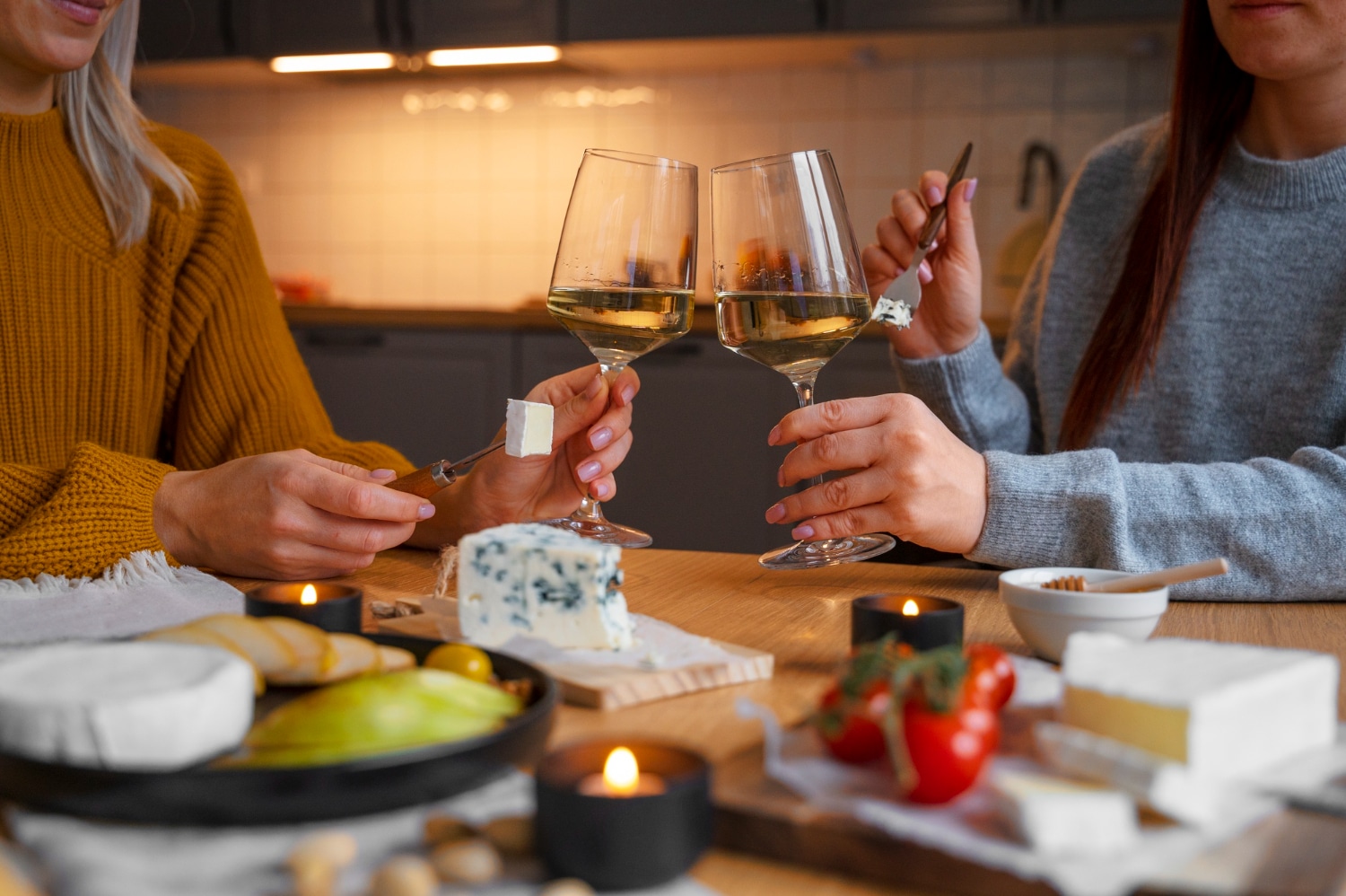 Two women toast with glasses of white wine while enjoying a cheese platter, highlighting a cozy gathering.