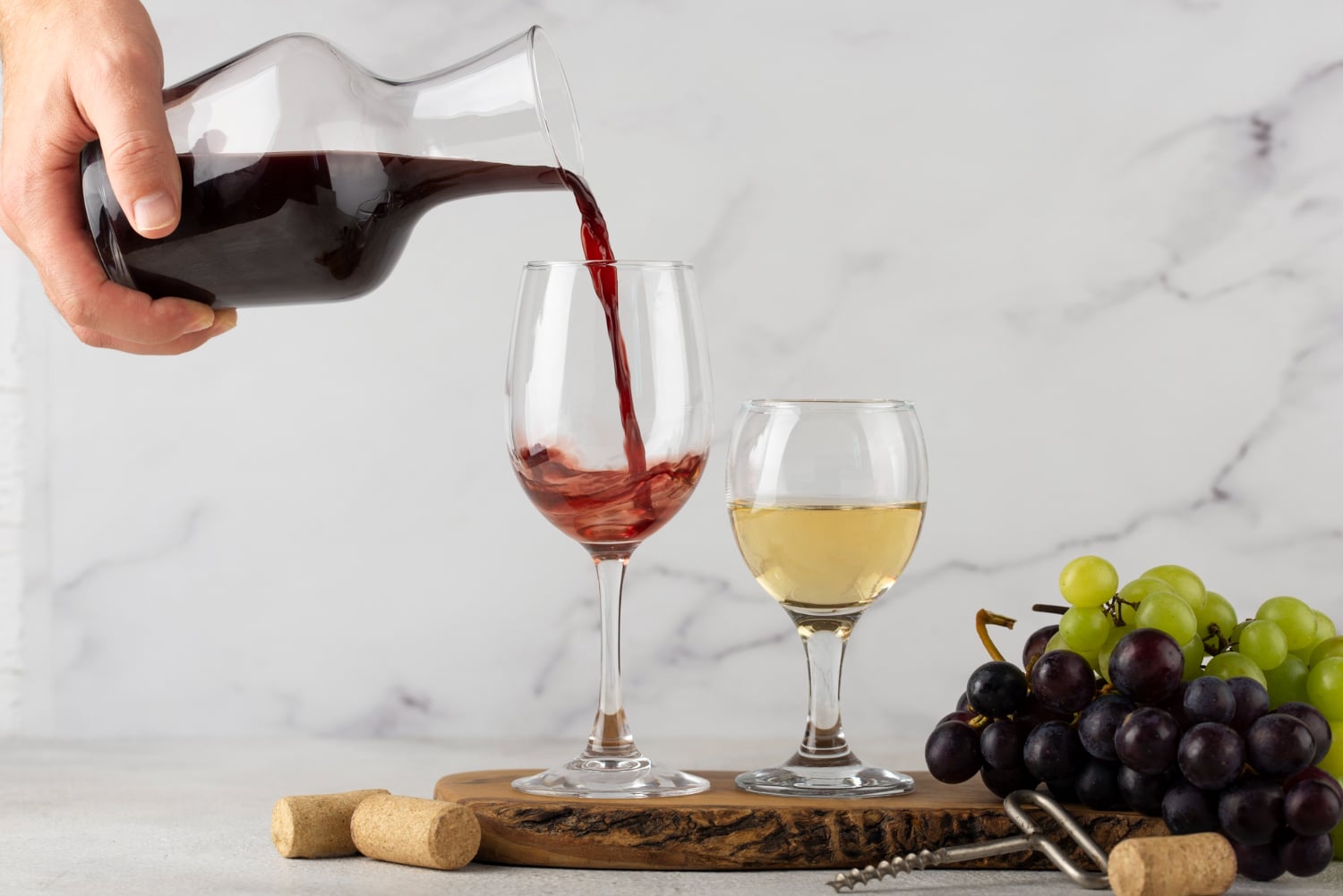 A hand pours red wine from a carafe into a glass, beside a white wine glass and a bunch of grapes on a wooden board.